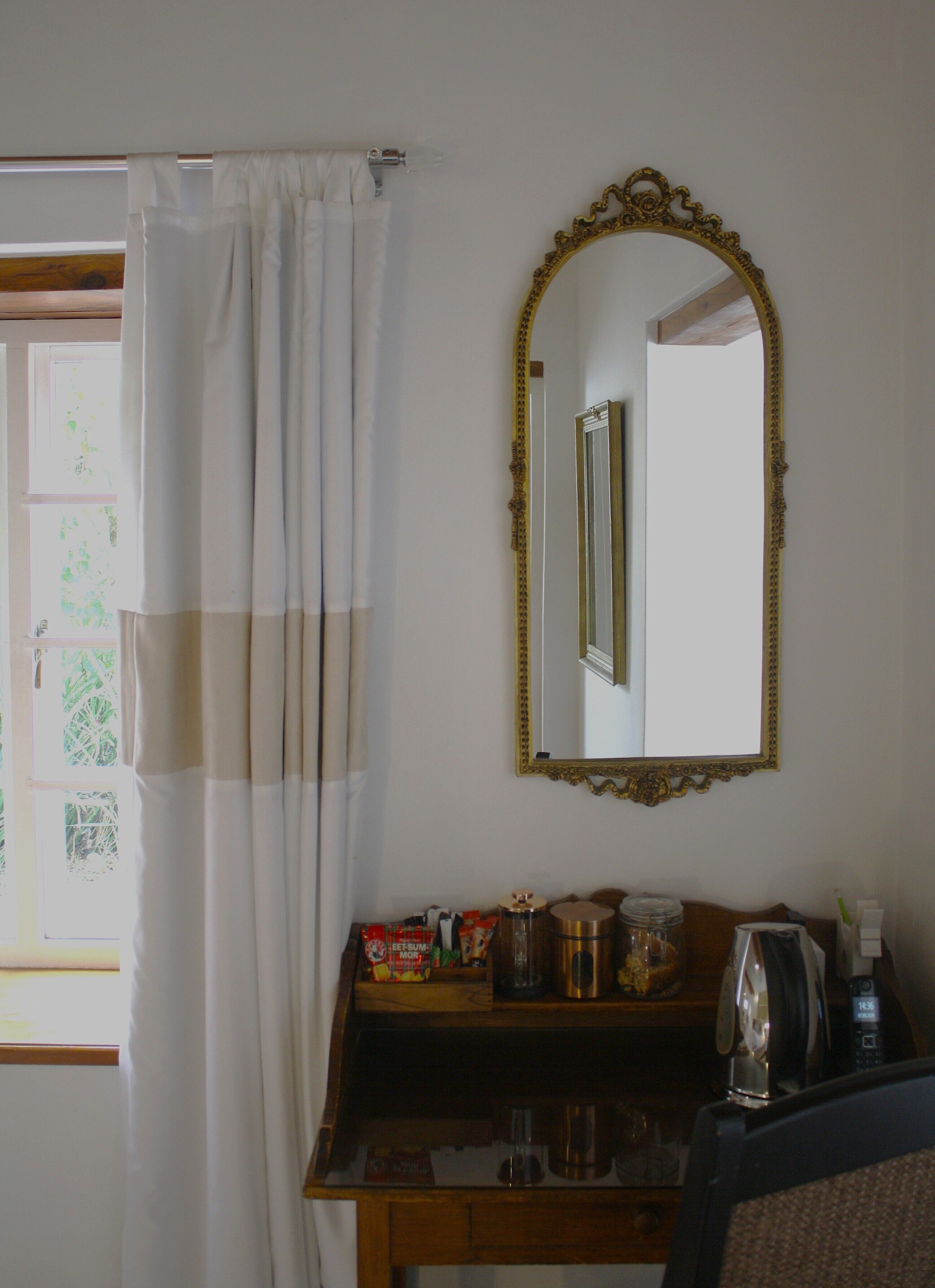 A bathroom with a wooden vanity, a mirror with an ornate frame, a shower curtain, and a window with a view of greenery.
