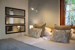 A neatly made bed with a grey blanket, two pillows with a marble pattern, and a nightstand with a lamp. There are three framed mirrors on the wall and a window with wooden shutters.