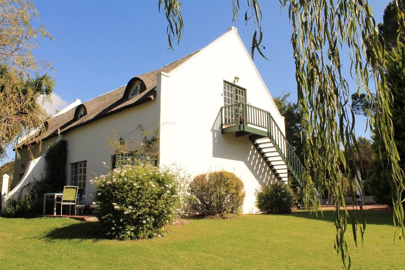 A white building with a steeply pitched roof, a staircase leading to a balcony, and a small garden area with a chair and table.