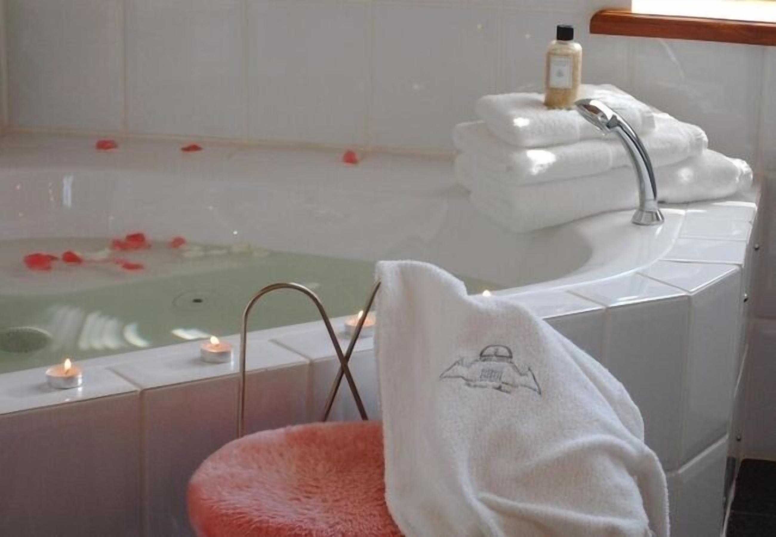 Ein Badezimmer mit Badewanne, Waschbecken und einem rosa Sessel. Auf dem Rand der Badewanne stehen Kerzen und auf dem Sessel liegt ein Handtuch.