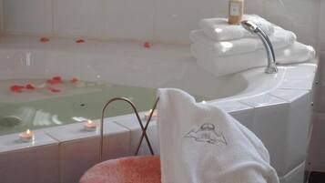 Ein Badezimmer mit Badewanne, Waschbecken und einem rosa Sessel. Auf dem Rand der Badewanne stehen Kerzen und auf dem Sessel liegt ein Handtuch.