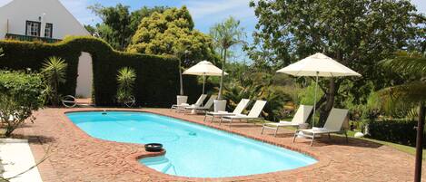 Una piscina con borde de ladrillo, rodeada de sillones blancos y sombrillas.