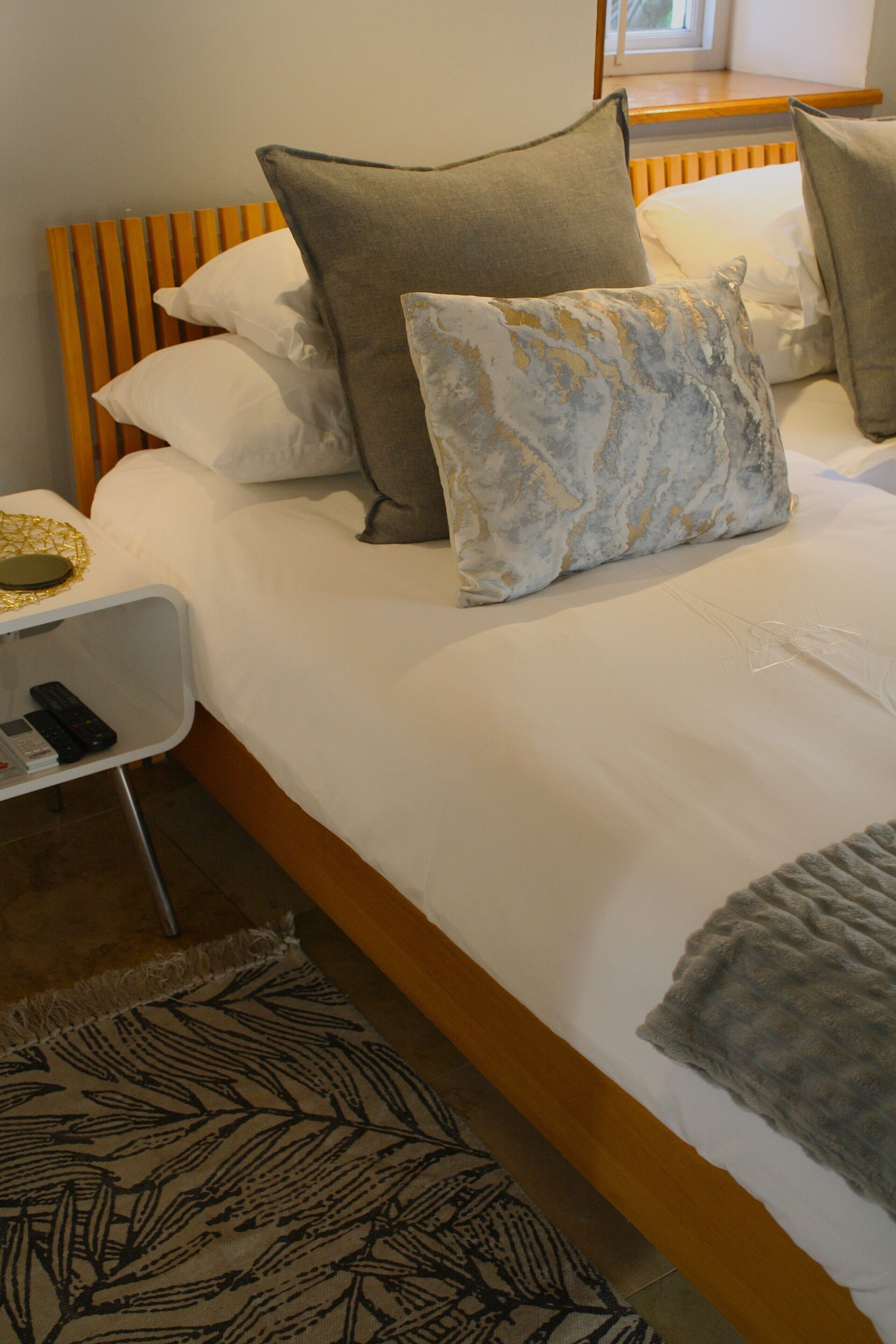Una cama bien hecha con ropa blanca y varios almohadones, una mesita de noche con control remoto y una alfombra estampada en el suelo.