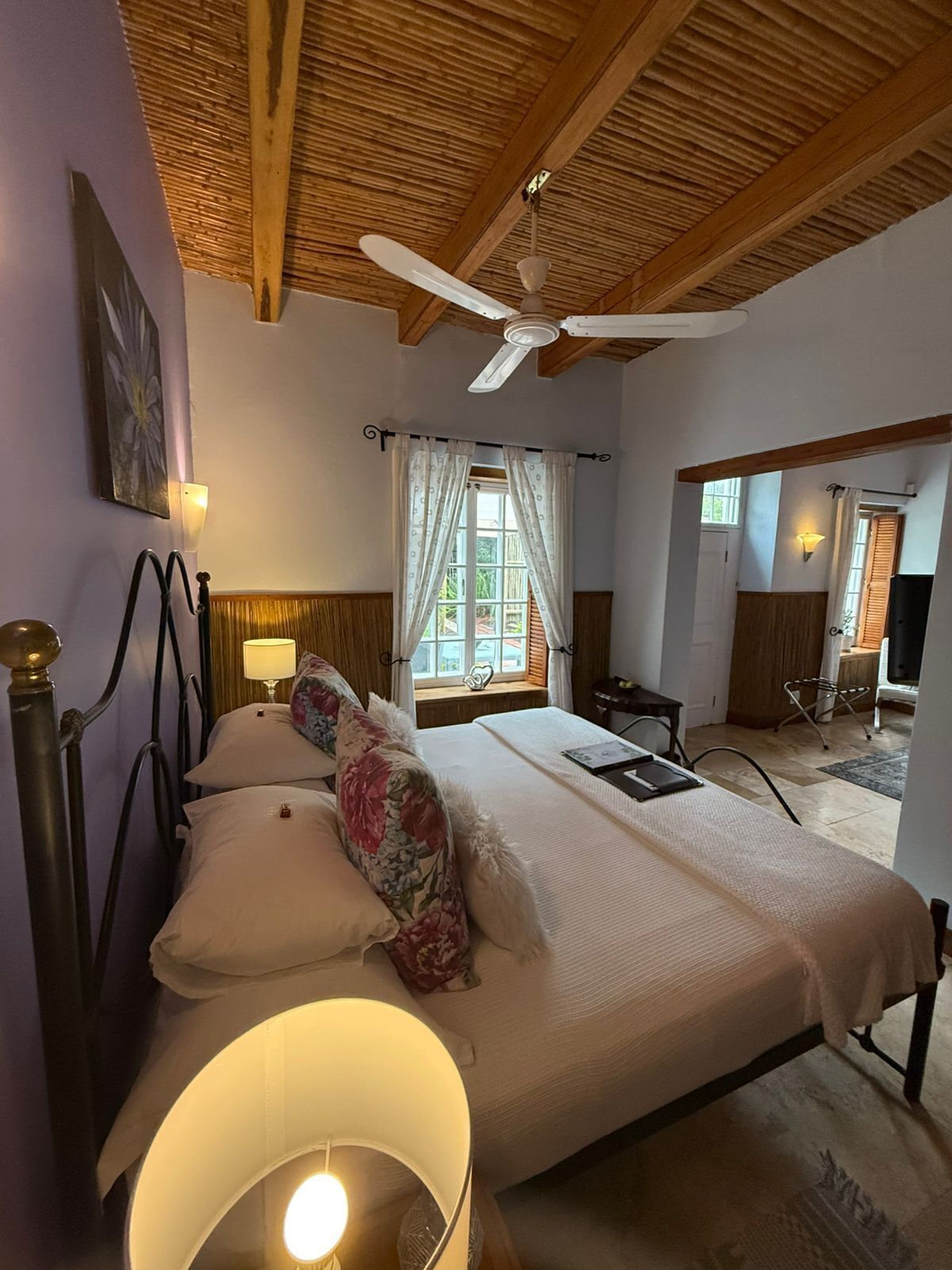 Une chambre à coucher avec un lit, un ventilateur de plafond, une fenêtre avec des rideaux et une table de chevet avec une lampe.
