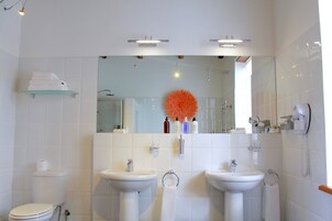 Un baño moderno con dos lavamanos, un inodoro y un estante de vidrio con toallas.