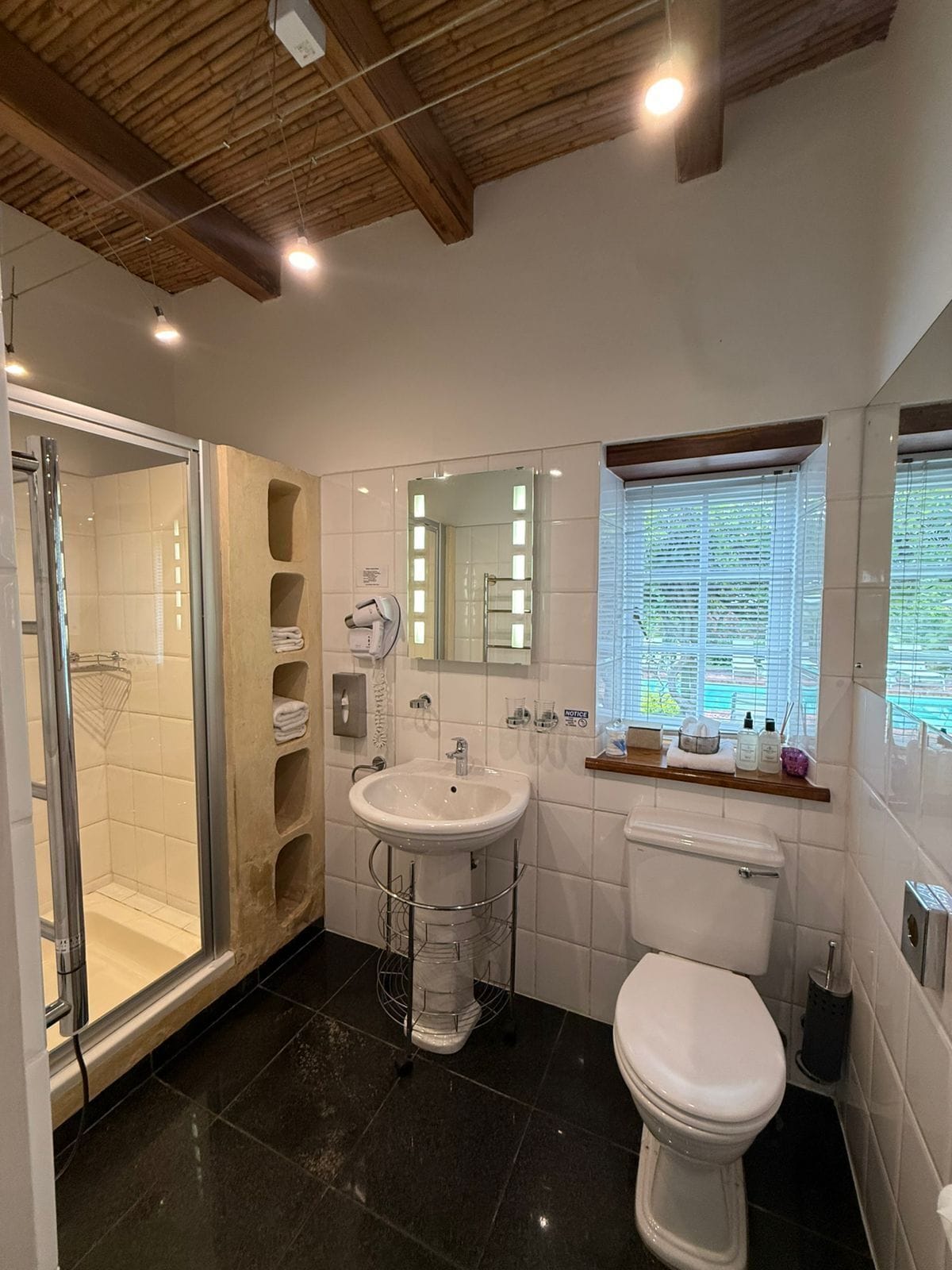 Et moderne badeværelse med brusekabine i glas, et væghængt håndvaskarrangement, et toilet og et vindue med persienner.