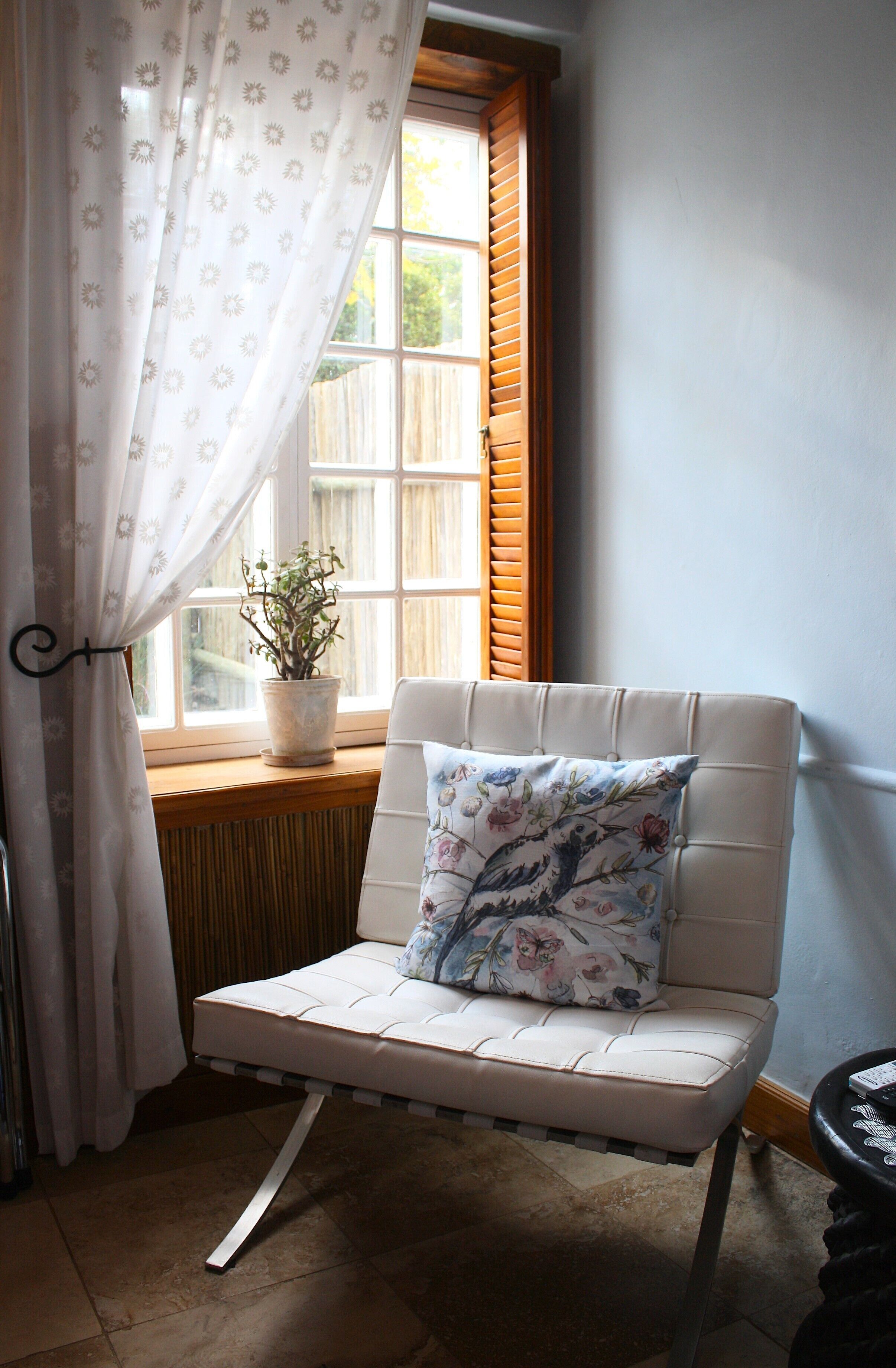 A room with a white cushioned chair with a floral pillow, a potted plant on a windowsill, and a window with wooden shutters.