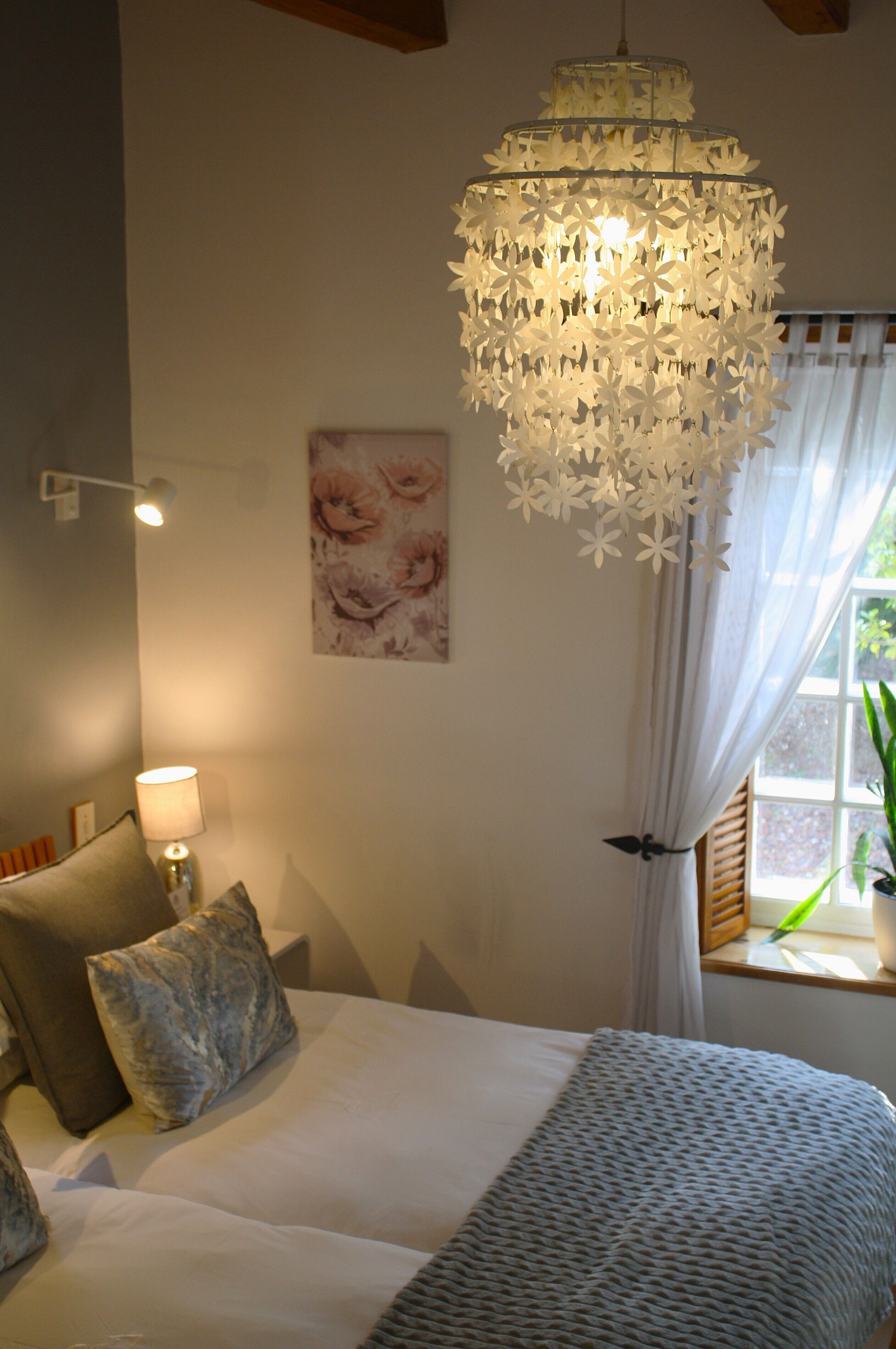 A bedroom with a bed, pillows, a wall-mounted lamp, a decorative chandelier, and a window with curtains.