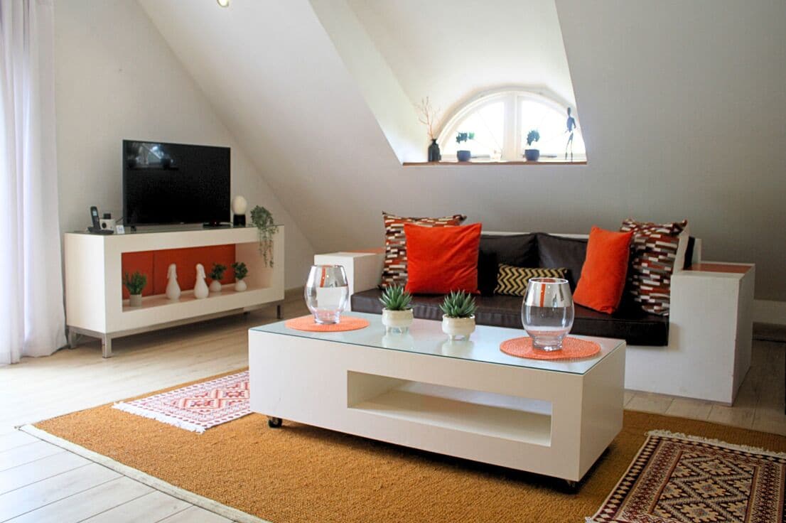 A modern living room with a glass coffee table, a sofa with orange cushions, a white entertainment unit, and a small window with plants.