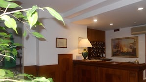 A reception area with a wooden desk, a green lamp, framed artwork, and a wall-mounted air conditioning unit. - Hotel Traghetto (Civitavecchia)