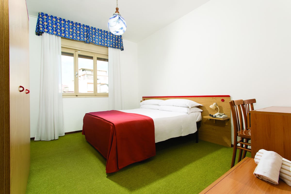 A hotel room with a bed, a wooden nightstand, a desk, and a green carpet.