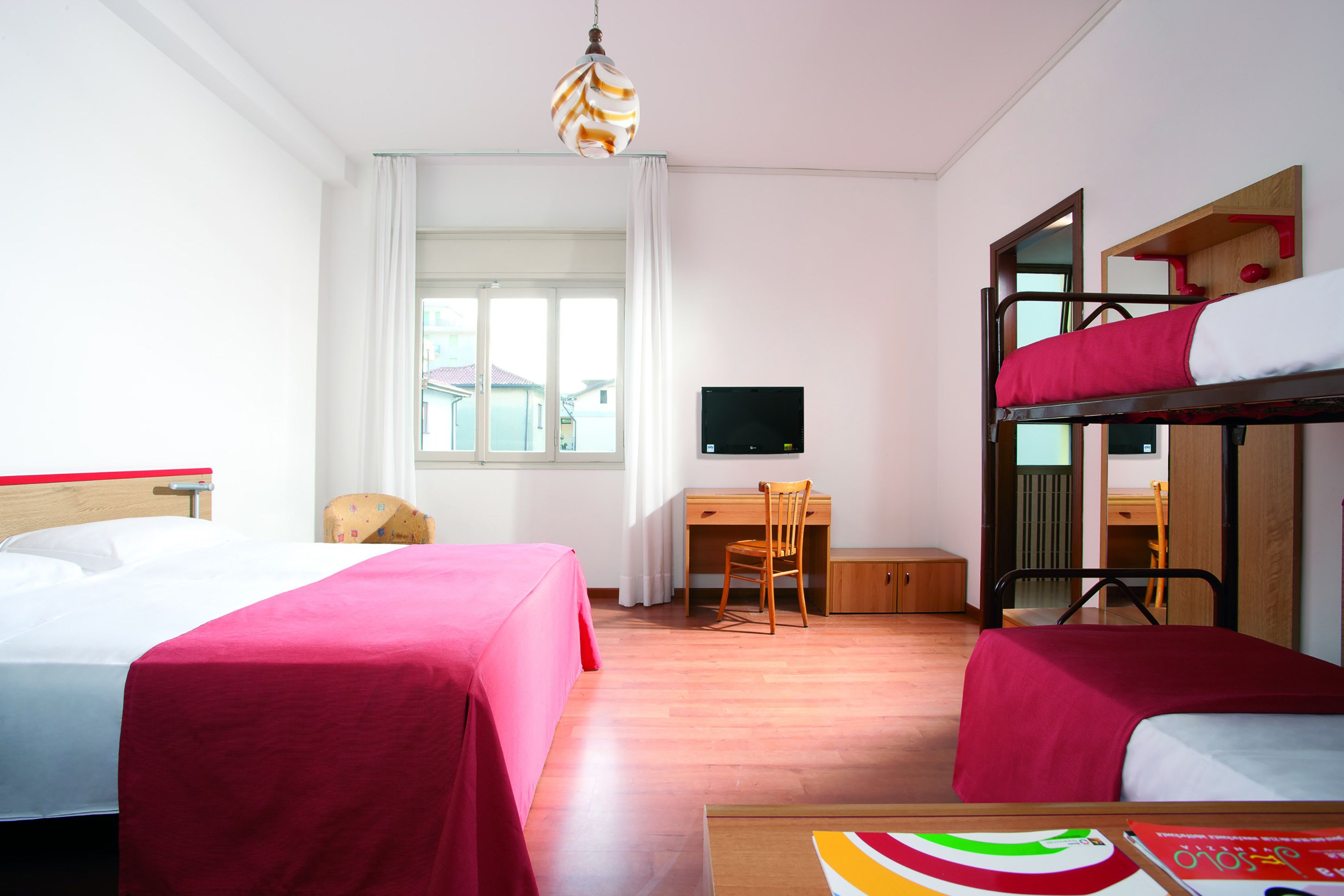 Quarto de hotel com duas camas, uma escrivaninha, uma cadeira, uma televisão e uma cama beliche.