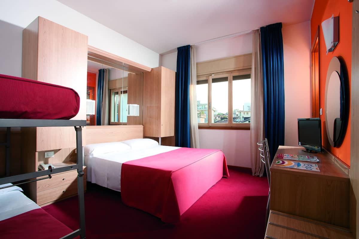 A hotel room with a bunk bed, a single bed, a desk with a computer, a mirror, and a window with curtains.