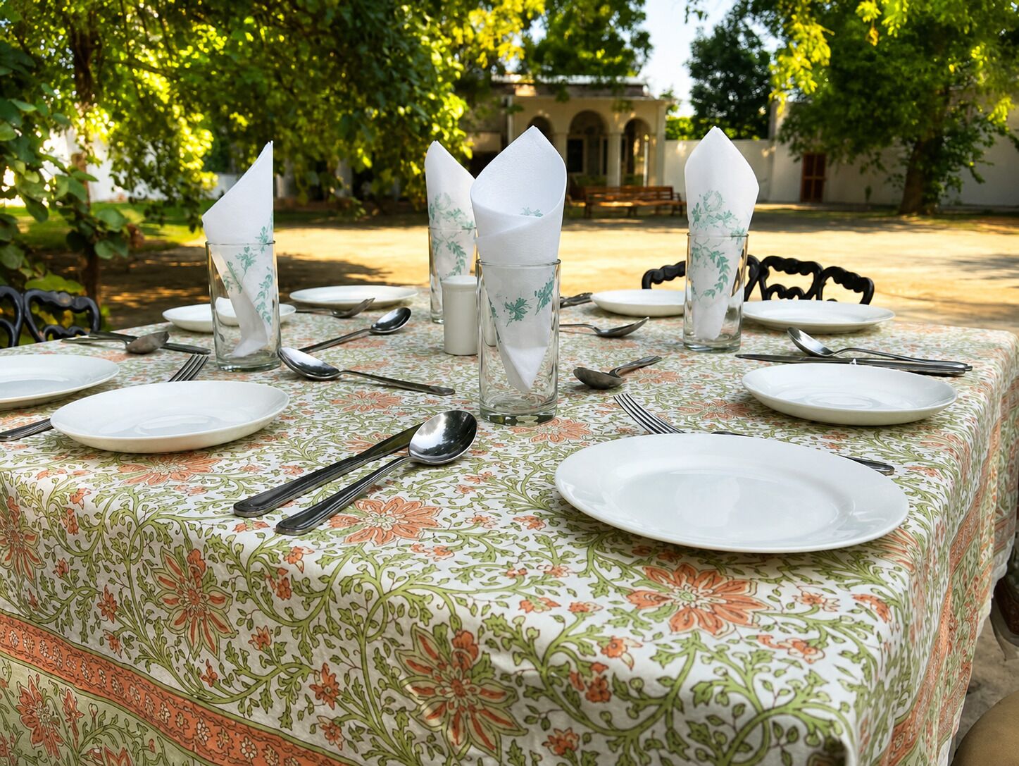 A table set outdoors with plates, cutlery, and glasses, surrounded by trees and a building in the background.