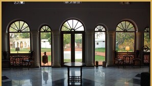A spacious lobby with a polished floor, a central table, and arched windows offering views of a courtyard with a building and greenery.