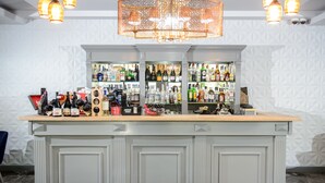 Uma área de bar com balcão de madeira, prateleiras com diversas garrafas de bebidas e um lustre decorativo.