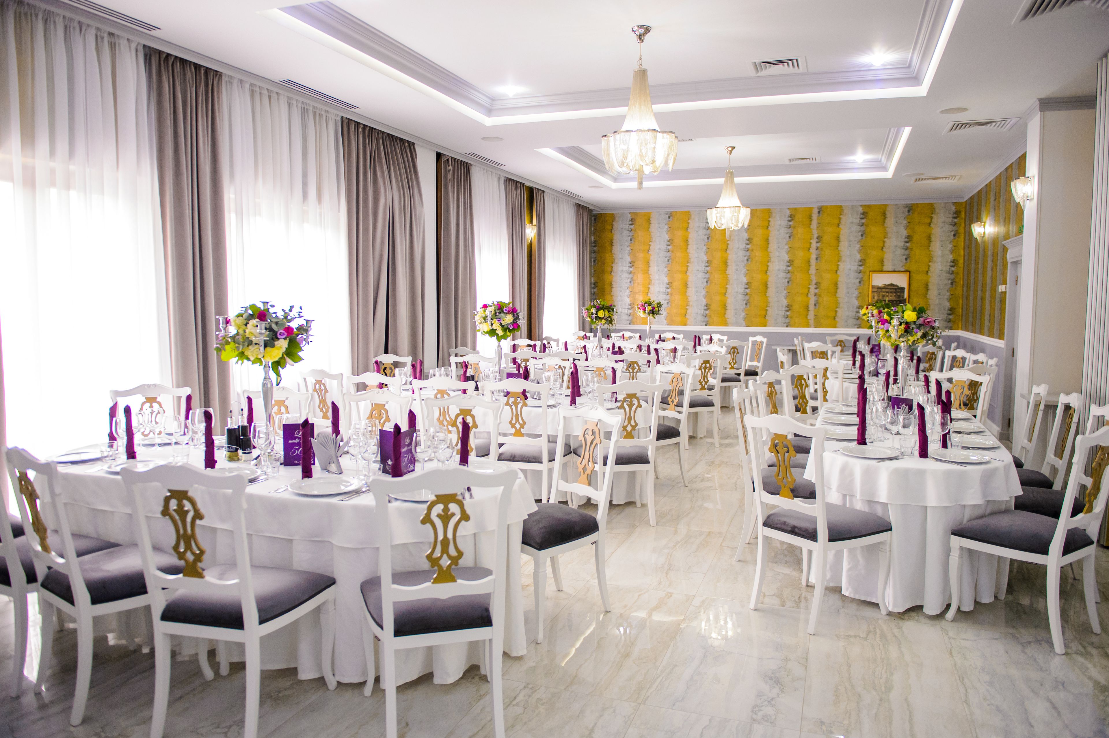 A banquet hall with round tables, white chairs, and chandeliers.