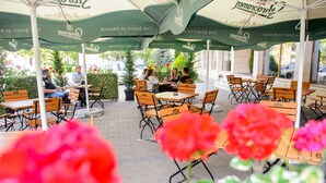 Café ao ar livre com mesas e cadeiras sob guarda-sóis, rodeado por vegetação e flores.