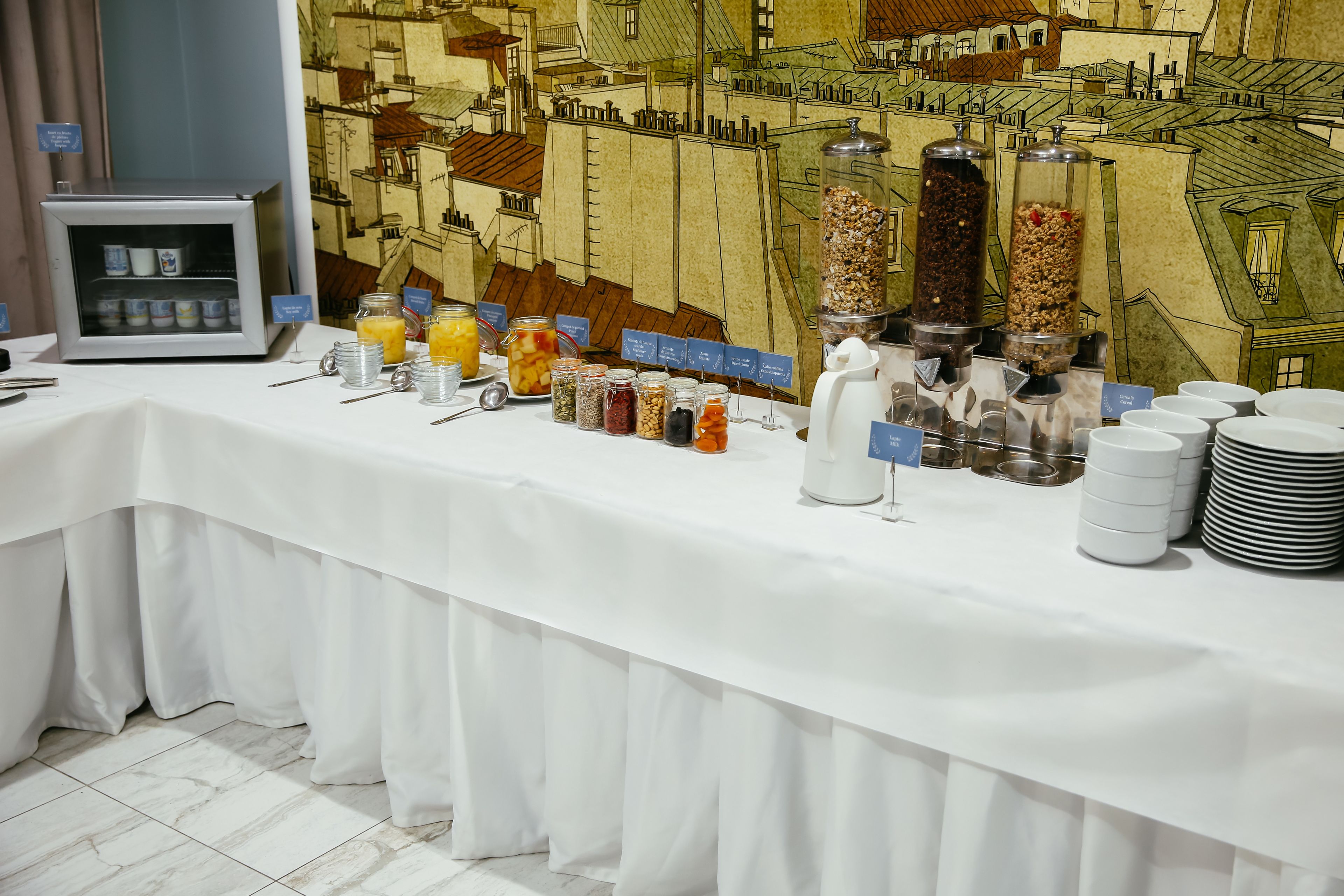 A breakfast buffet with various food items and beverages, a coffee station, and a backdrop of a cityscape mural.