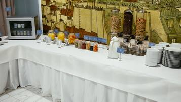 A breakfast buffet with various food items and beverages, a coffee station, and a backdrop of a cityscape mural.
