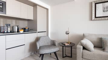 A modern kitchen with white cabinets, a coffee machine, and a kettle. There is a grey armchair and a small round table with a lamp.