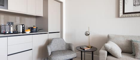 Eine moderne Küche mit weißen Schränken, einer Kaffeemaschine und einem Wasserkocher. Ein grauer Sessel und ein kleiner runder Tisch mit einer Lampe stehen dort.