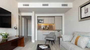 A modern living room with a flat-screen TV, a sofa, a coffee table, and a small side table. There is a kitchen area with a microwave and cabinets.