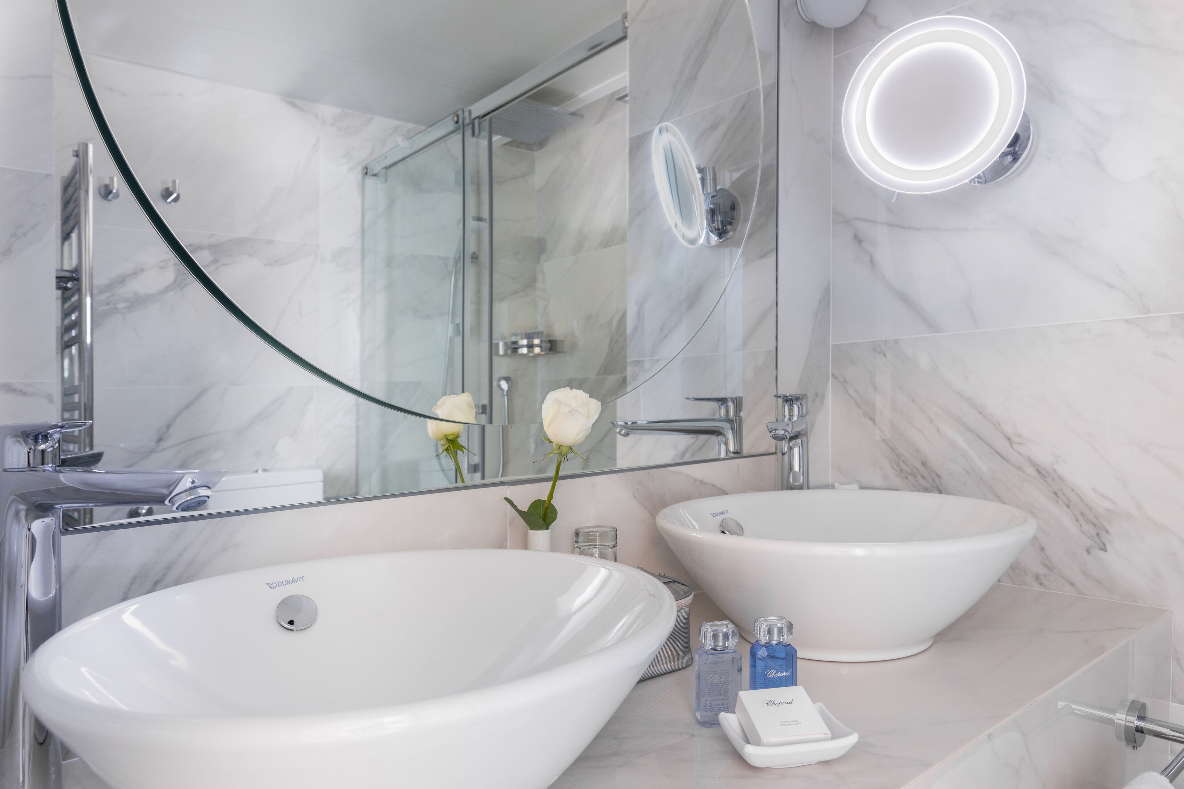 A modern bathroom with two white sinks, a glass shower enclosure, and a large mirror with a lighted frame.