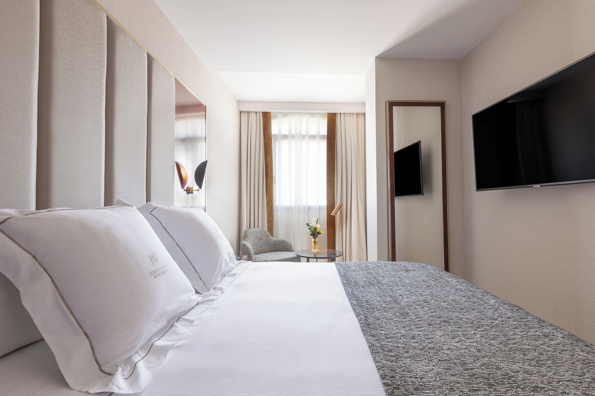A hotel room with a large bed, a TV mounted on the wall, a small table with a vase of flowers, and a chair.