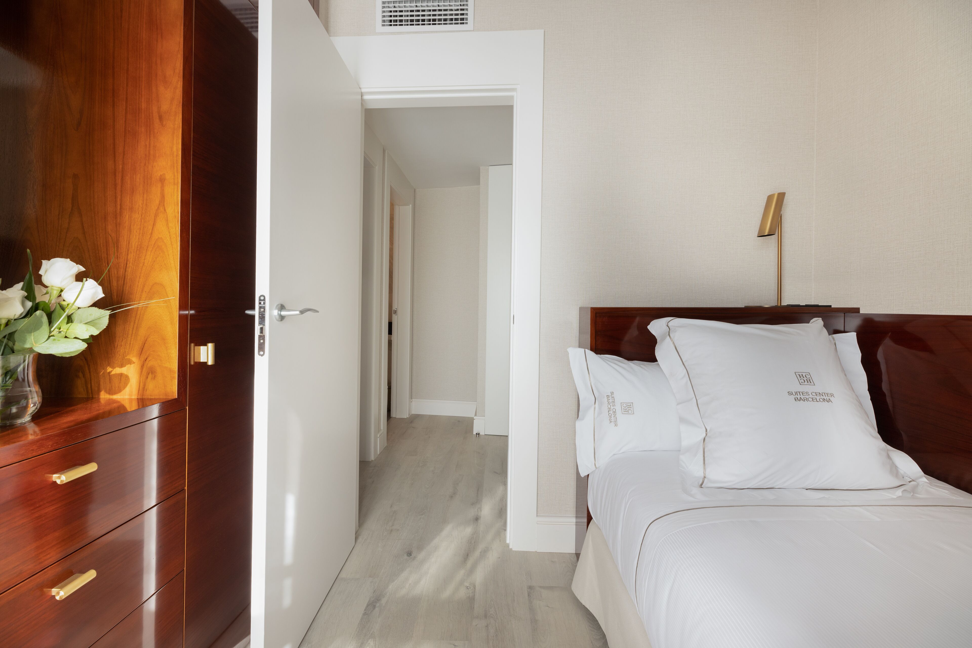 A hotel room with a wooden wardrobe, a bed with white bedding, and a bedside table with a lamp.