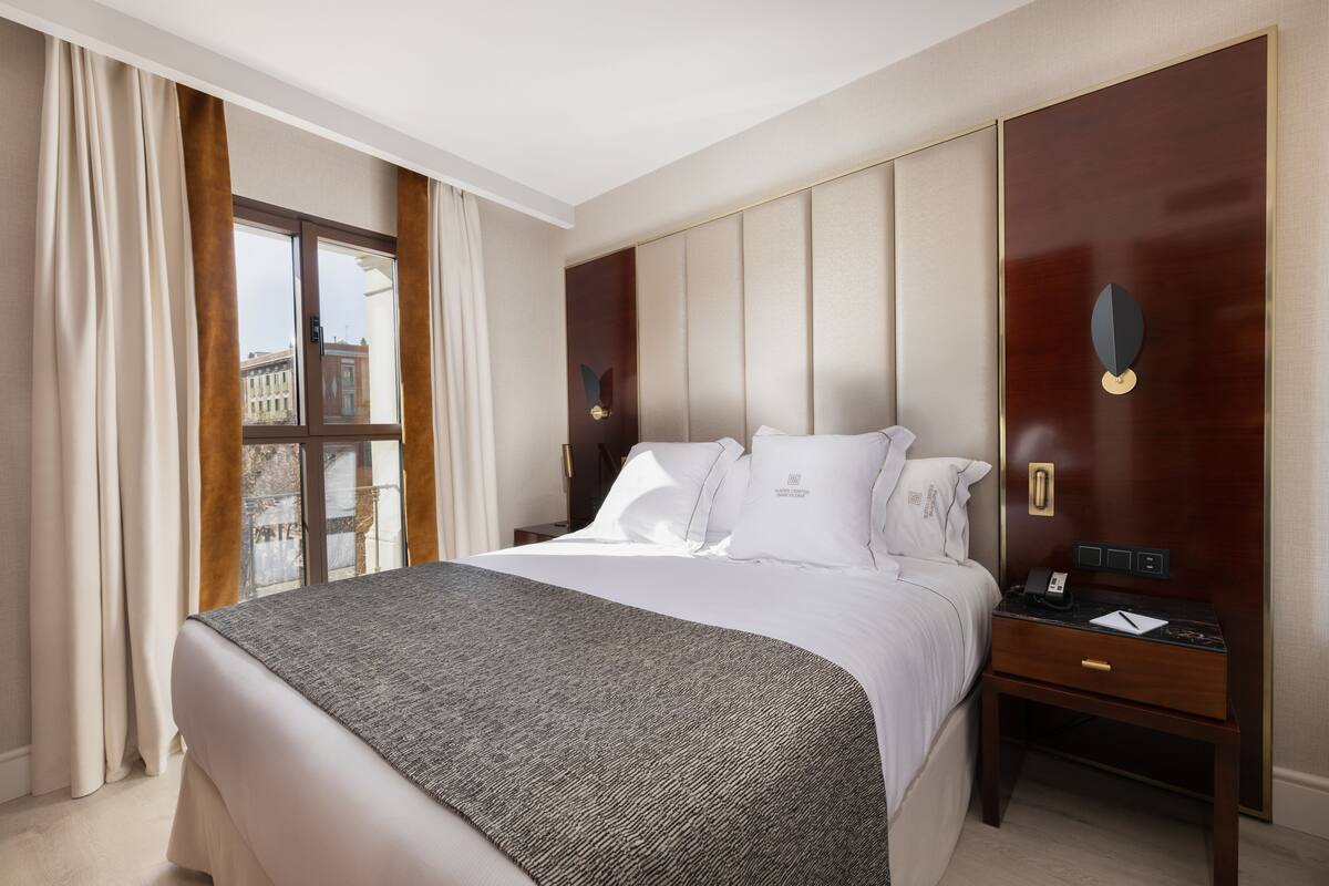 A hotel room with a large bed, a nightstand, a window with curtains, and a view of buildings outside.