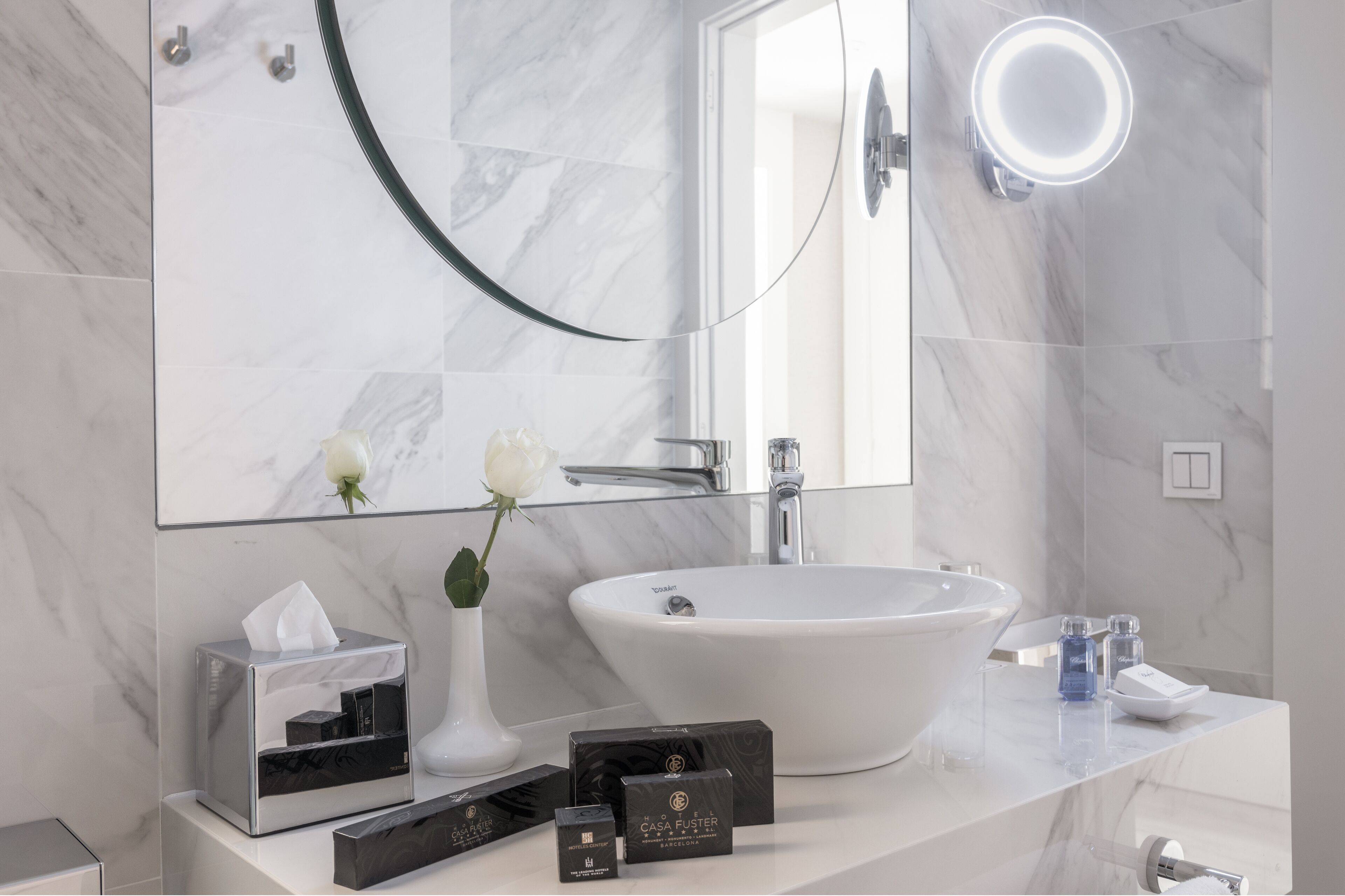 Un baño moderno con un lavamanos grande blanco, un florero de vidrio con una flor blanca y una caja de pañuelos en la encimera.