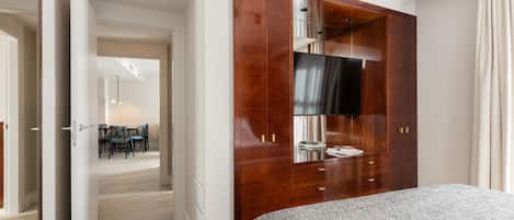 Habitación de hotel con una cama grande, un armario de madera con un televisor y un área de comedor visible a través de una puerta abierta.