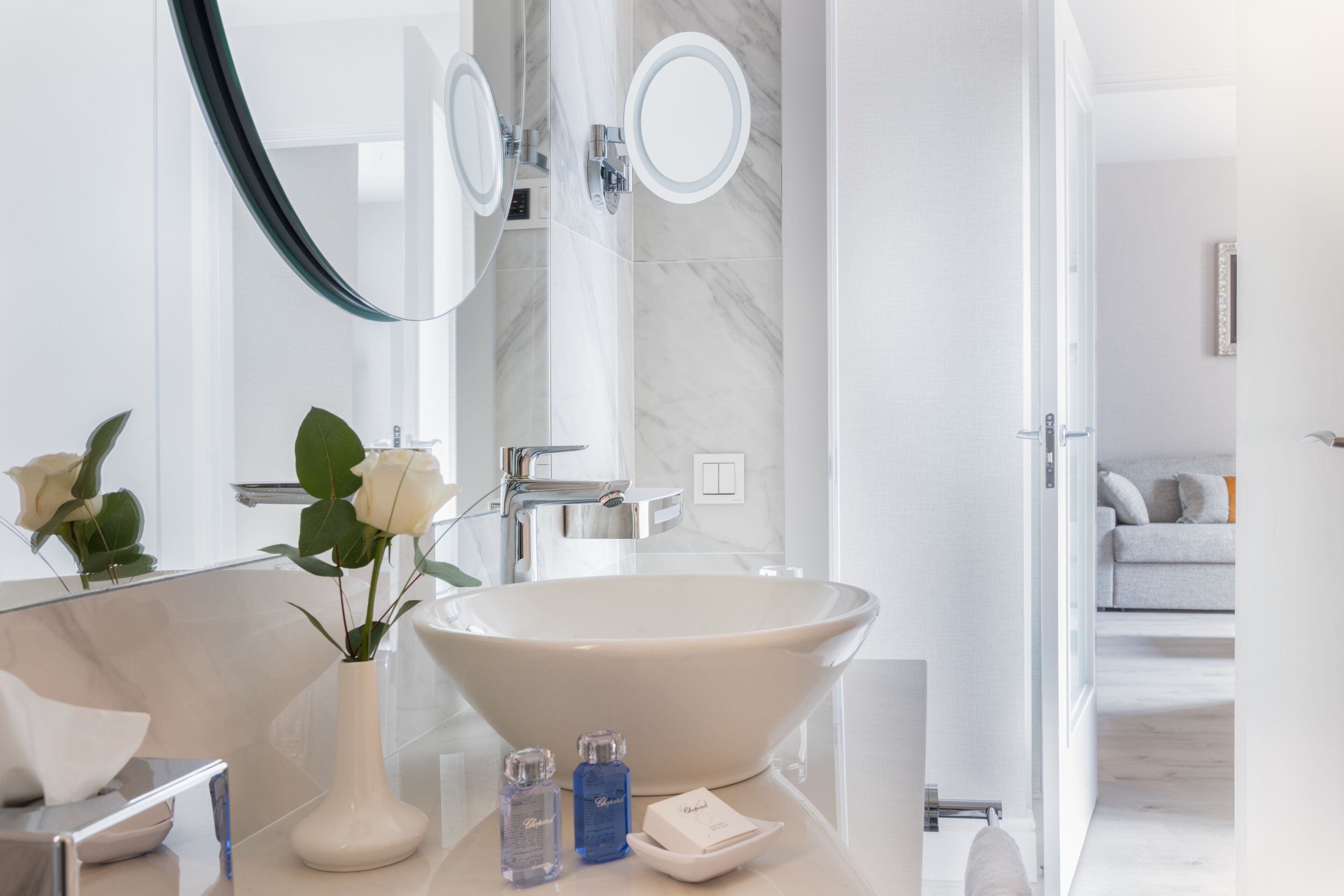 A modern bathroom with a large oval sink, a round mirror, and a vase with white flowers.