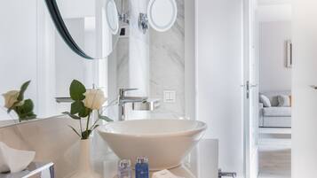 Un baño moderno con un lavabo ovalado grande, un espejo redondo y un florero con flores blancas.