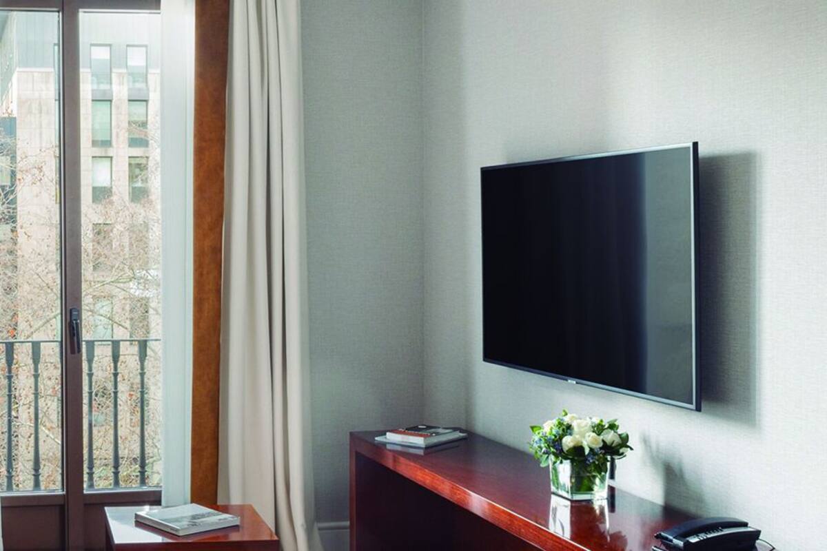 A modern hotel room with a sofa, a flat-screen TV, a desk with a vase of flowers, and a view of the outside balcony.
