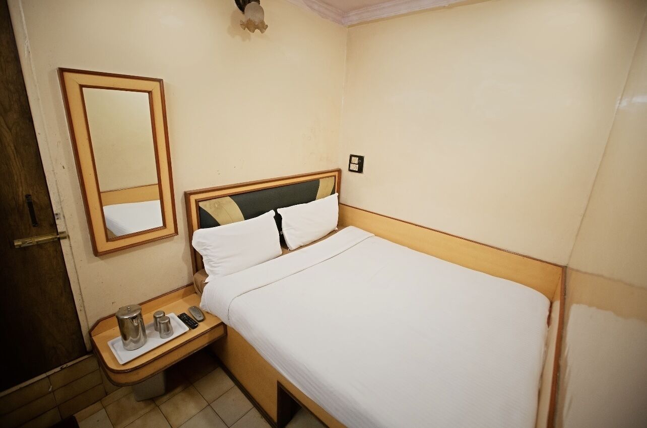 Habitación de hotel con una cama individual, una mesita de noche con lámpara, un espejo y una pequeña mesa con hervidor y control remoto.