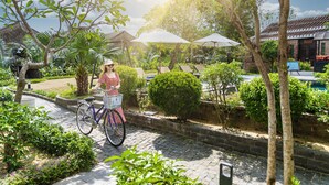 A person riding a bicycle with a basket, surrounded by lush greenery and a brick building with a thatched roof. - Hoi An Coco River Resort & Spa (Da Nang)