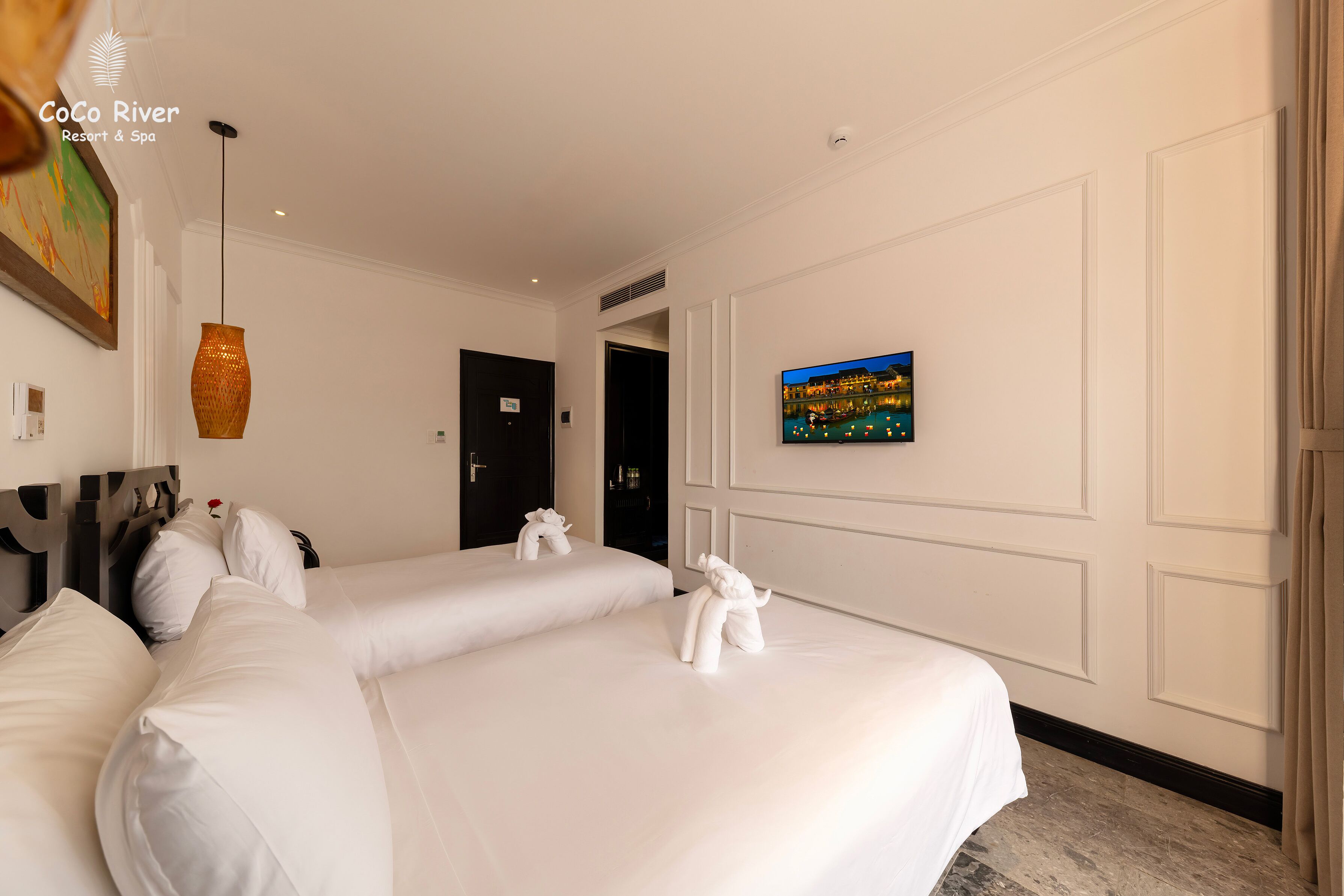 Habitación de hotel con dos camas, un televisor de pantalla plana montado en la pared y una figura decorativa de elefante en la cabecera.