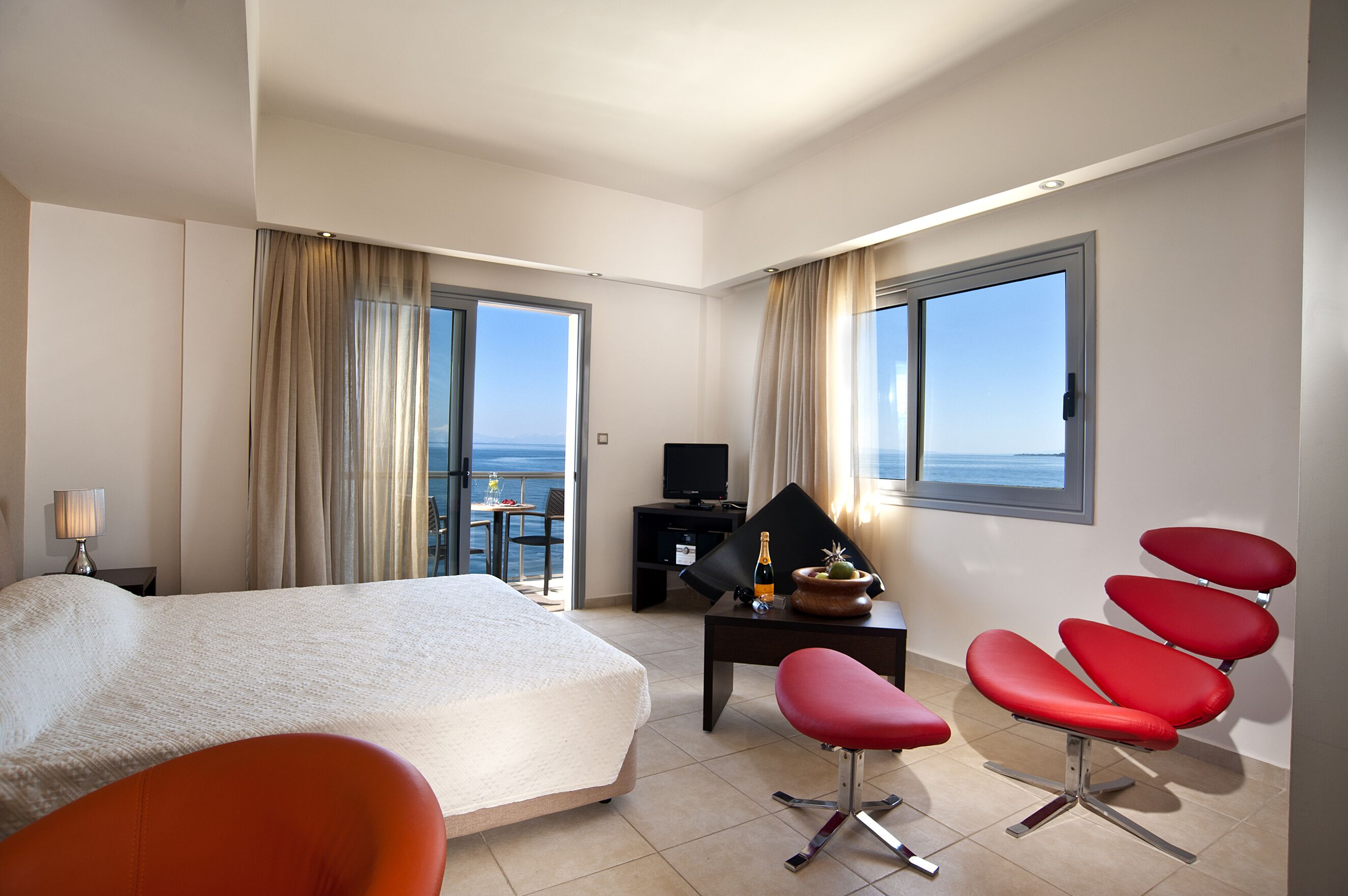 A hotel room with a bed, a desk, a TV, a chair, and a view of the sea.