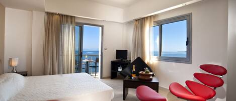 A hotel room with a bed, a desk, a TV, a chair, and a view of the sea.