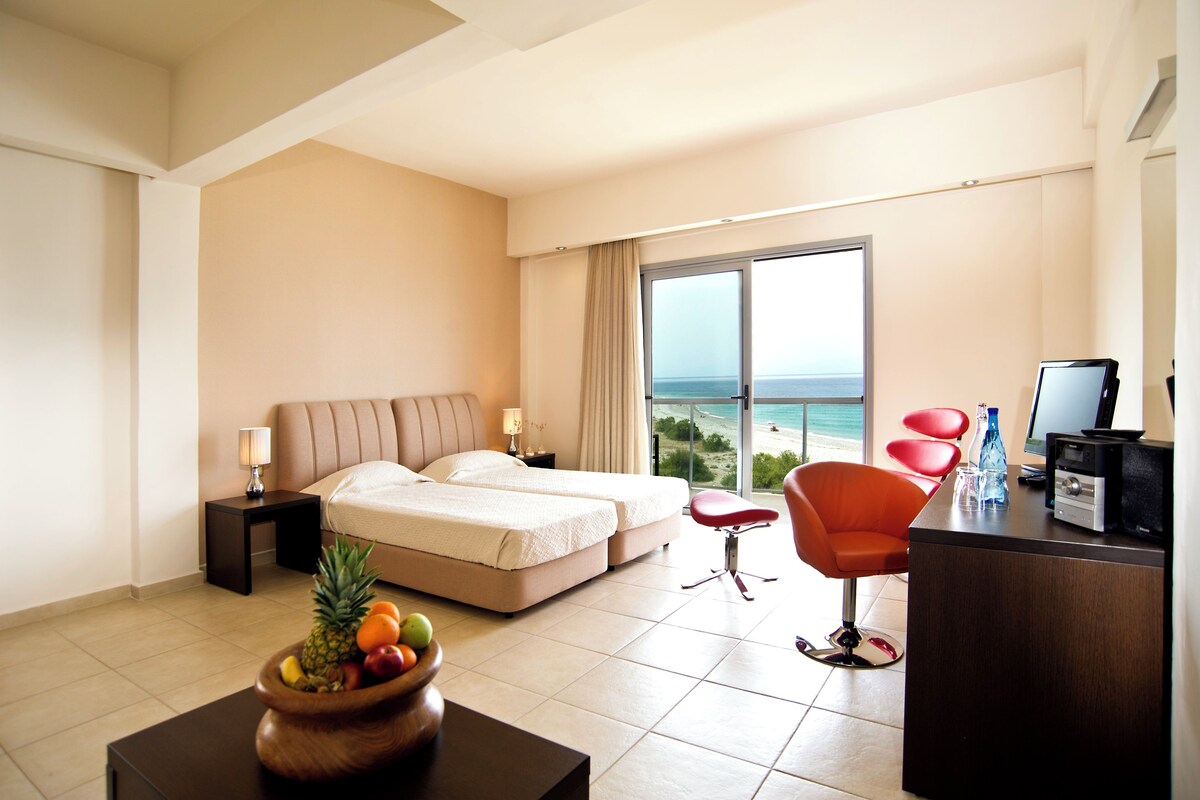 A hotel room with a bed, a desk with a computer, a chair, a fruit bowl, and a view of the beach.