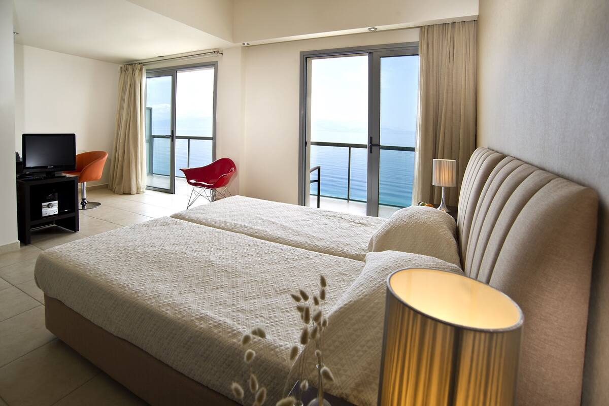 Ein Hotelzimmer mit einem großen Bett, einem Fernseher, einem roten Sessel und Meerblick durch die Fenster.