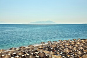 A beach with rows of sun loungers and thatched umbrellas, a clear blue sea, and distant mountains. - Sikyon Coast Hotel & Resort (Xylokastro-Evrostina)