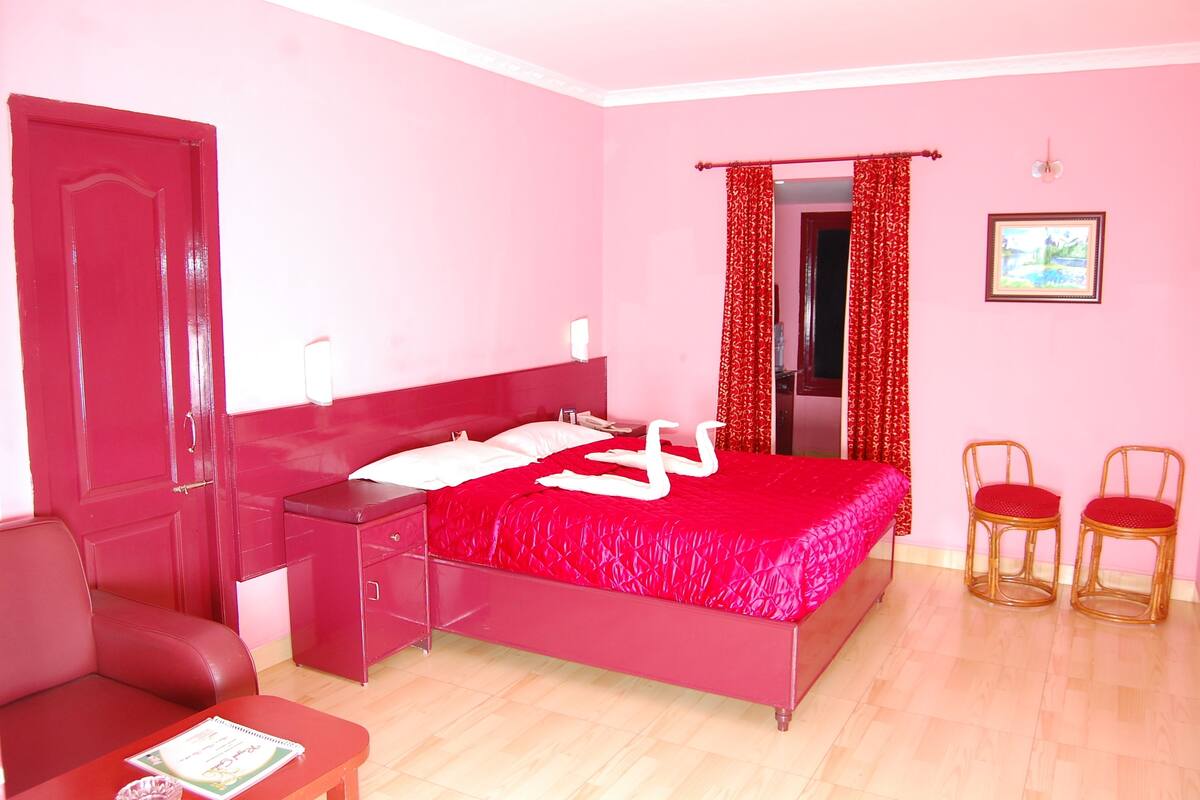 A room with a bed, a red door, a small table with a booklet, two wicker chairs, and a red curtain.