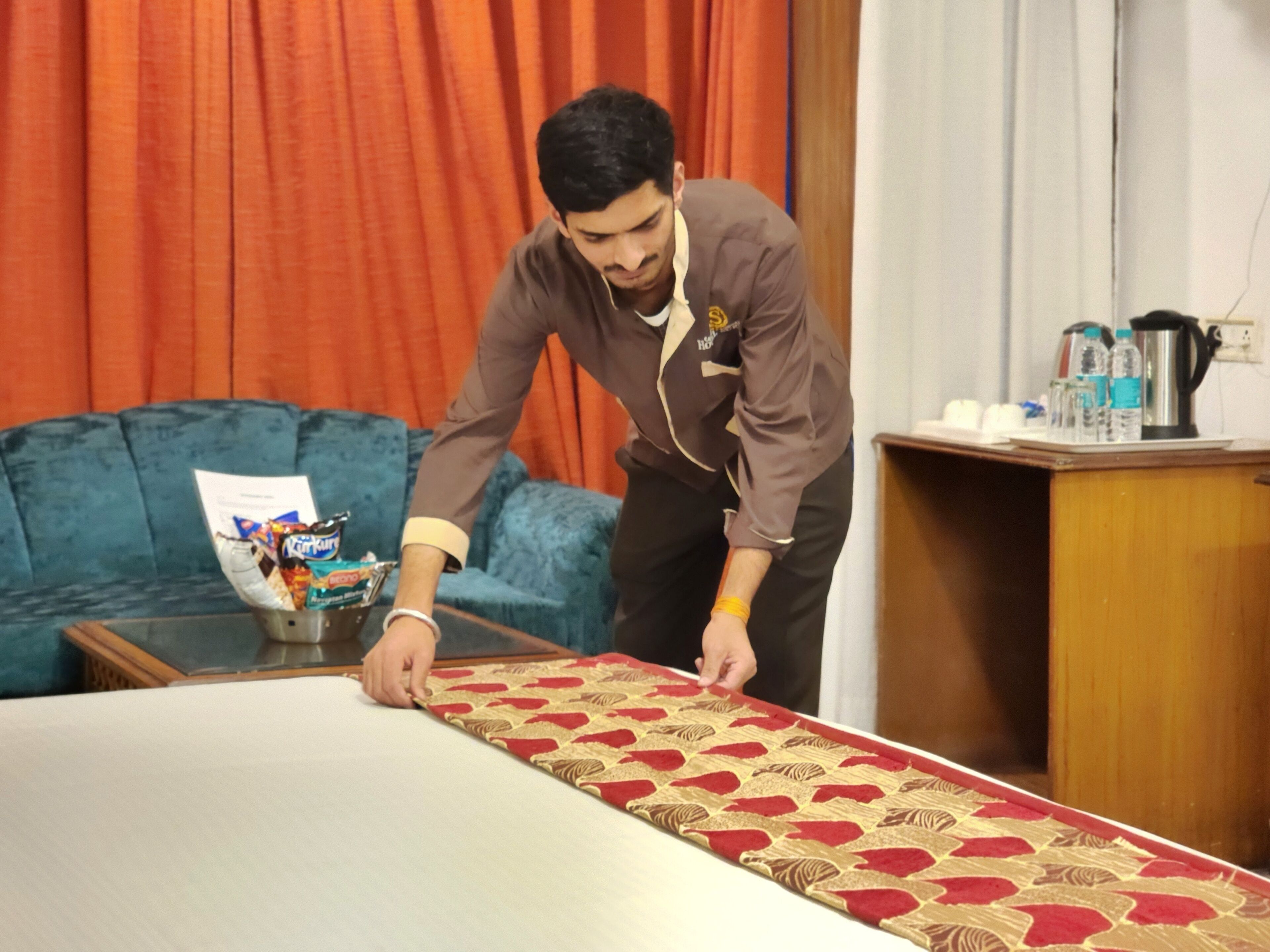 A person arranging a bed with a patterned bedspread in a room with a blue sofa and a wooden side table.