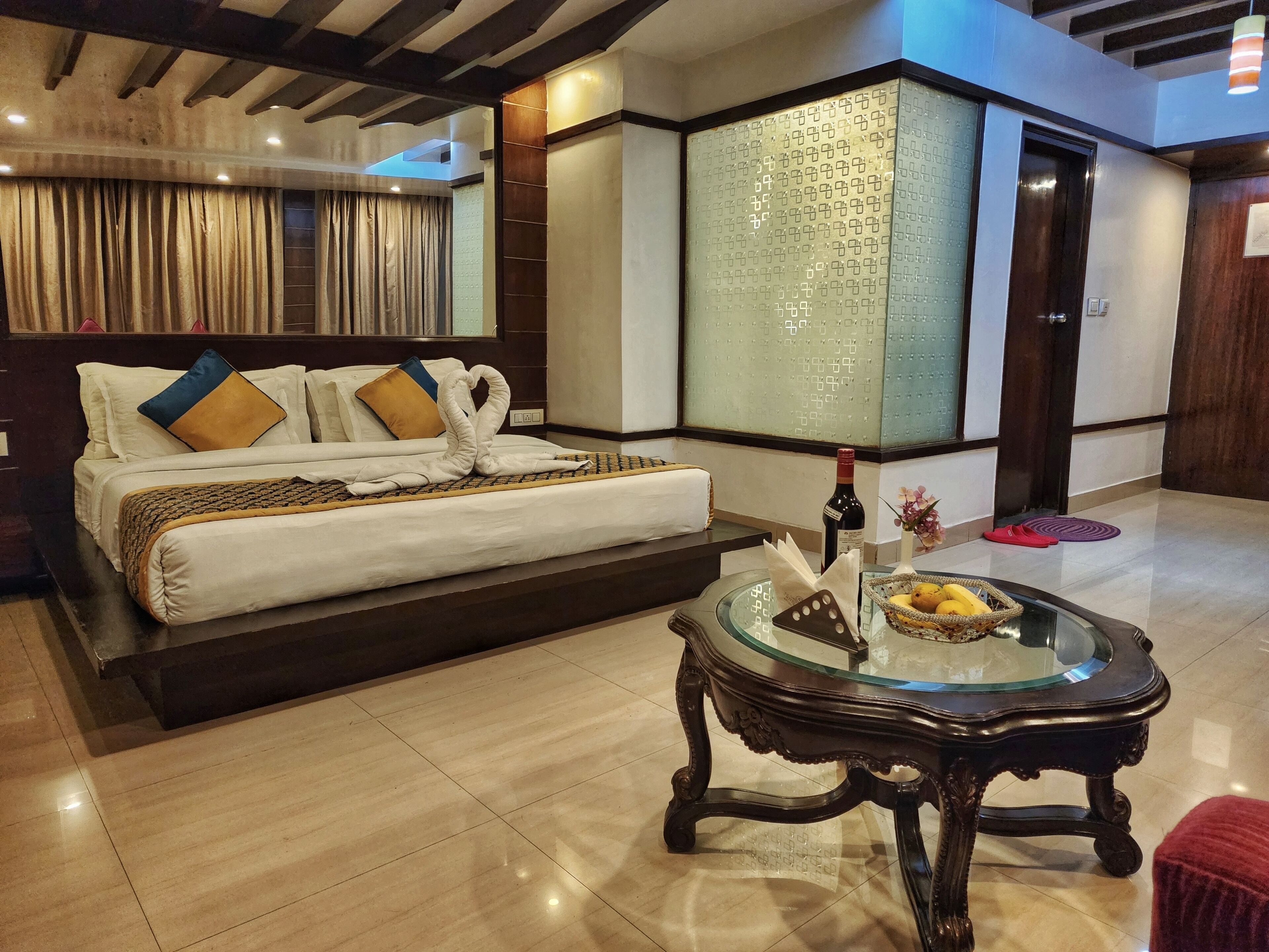 A hotel room with a bed, a glass-top table with a bottle and fruit, and a wooden floor.