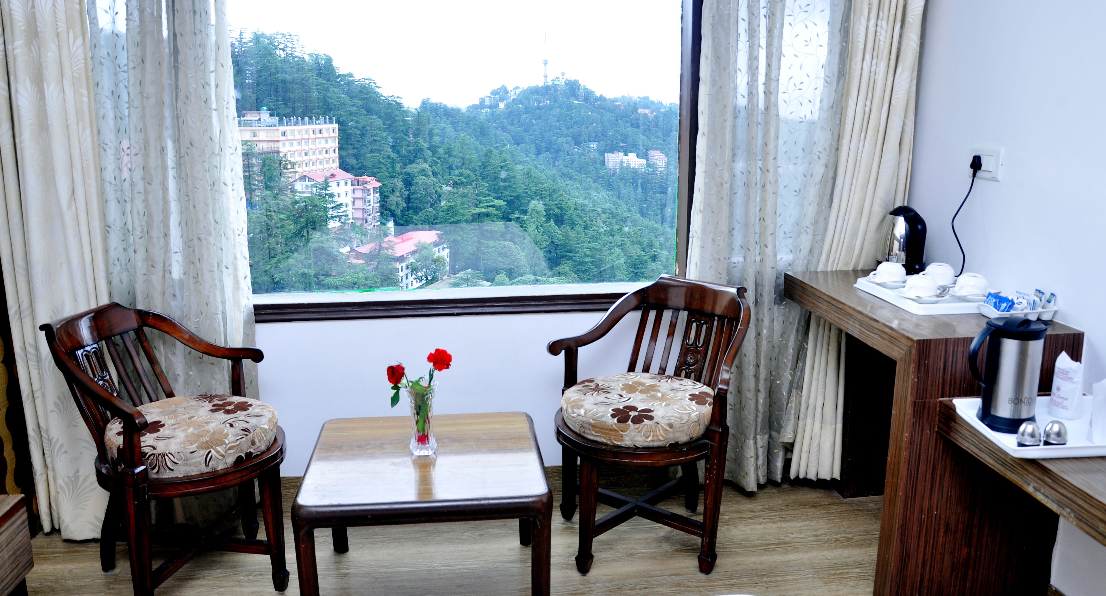 A room with two wooden chairs, a small table, a vase with flowers, and a view of a forested area through a window.
