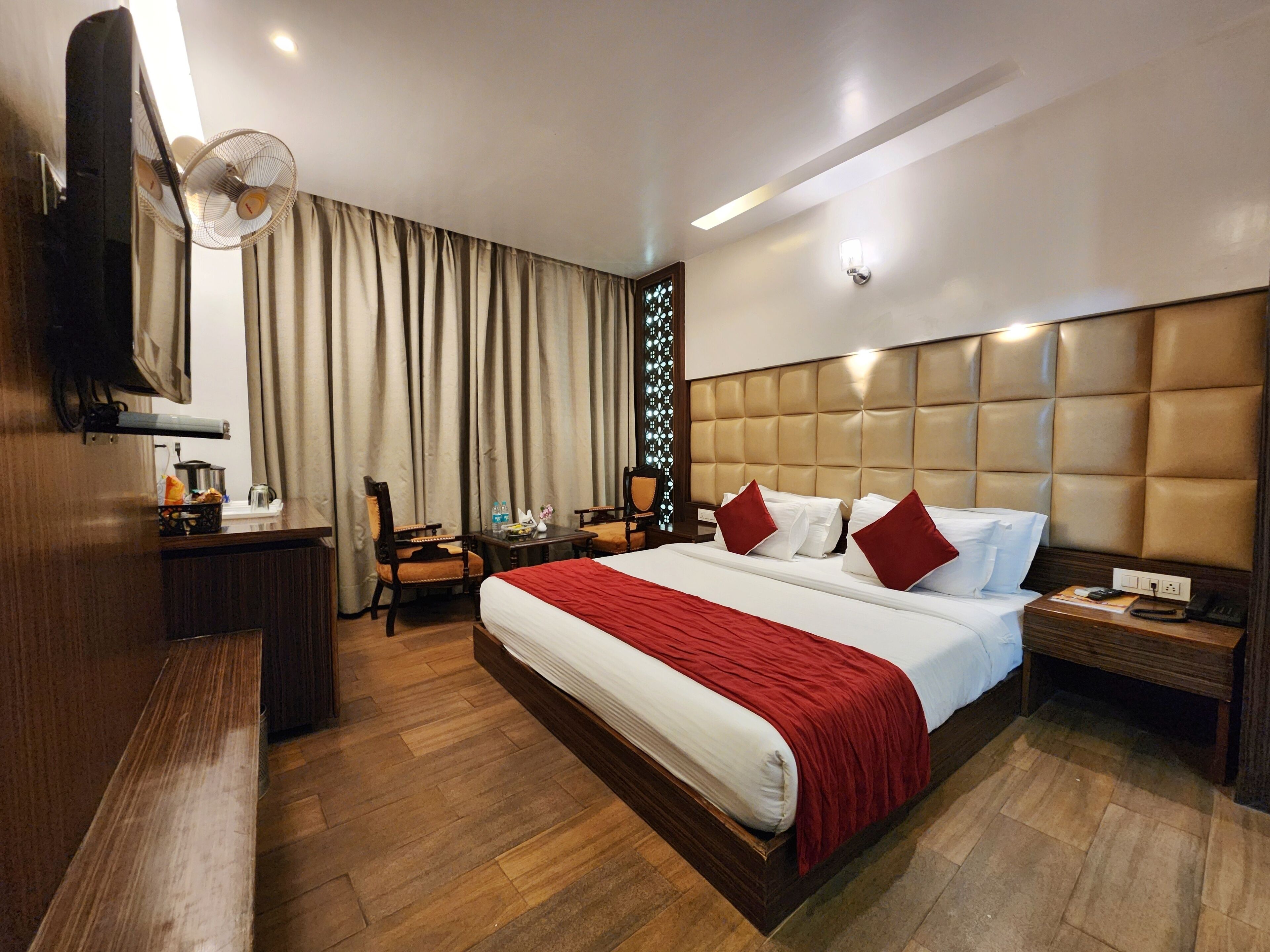 A hotel room with a large bed, a bedside table, a nightstand, a desk with a chair, a fan, and a window with curtains.
