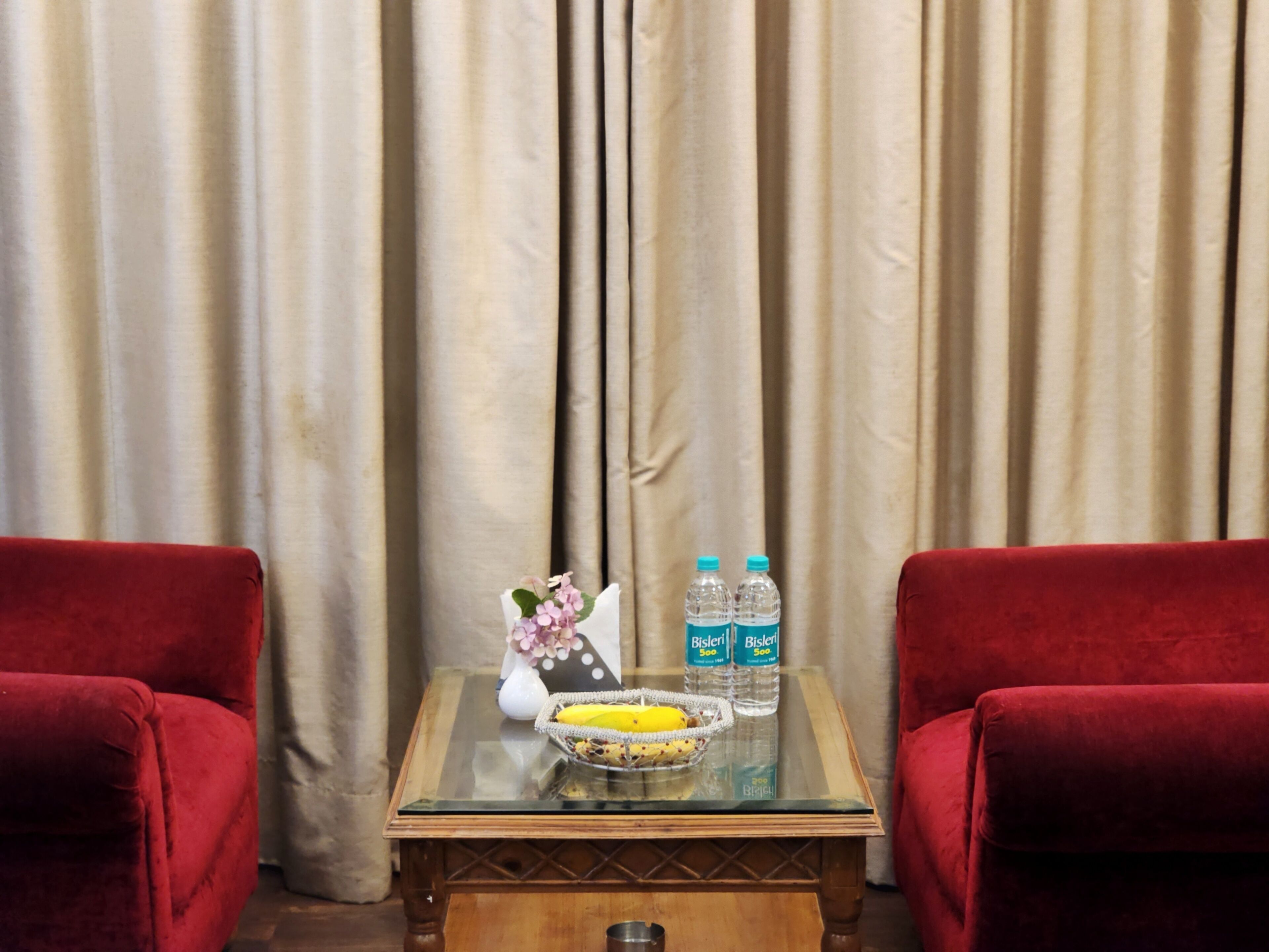Quarto com mesa de vidro, duas poltronas vermelhas, um vaso com flores e duas garrafas de água Bisleri.